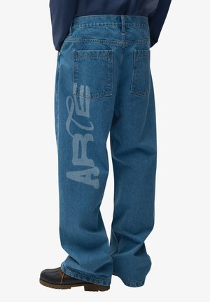 Persona che indossa jeans blu larghi con la parola "AFTER" stampata verticalmente sulla gamba sinistra e scarpe nere con suola marrone.