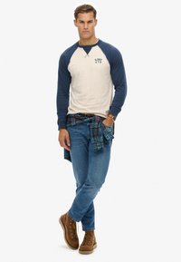 Chemise à manches longues en crème et marine, avec un petit logo. Associée à un jean bleu et des bottes en cuir marron à lacets. Chemise à carreaux nouée à la taille.