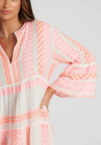 Lekka, biała sukienka z różowymi geometrycznymi wzorami, dekoltem w serek i dzwonowymi rękawami. Tkanina ma fakturowane wykończenie i warstwowy dół.