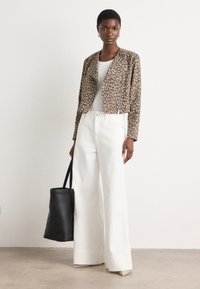 Jas met luipaardprint, cropped, beige en bruin, met zilveren ritsen. Witte wijde broek, effen, gecombineerd met een zwarte tote bag.