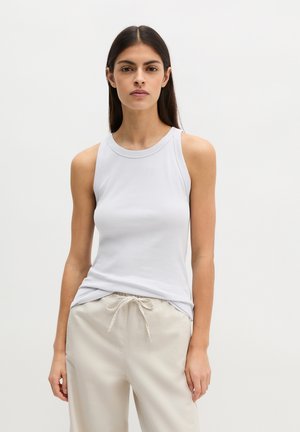 Kobieta o długich ciemnych włosach, ubrana w biały top bez rękawów i jasnobeżowe spodnie z sznurkiem, stojąca na tle jednolitego tła.