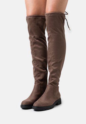 Hnědé semišové kozačky nad kolena s černou podrážkou, nošené na bosých nohou, s bočními zipy a stahovacími šňůrkami na horním zadním okraji.