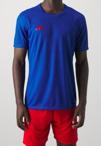 Blå idrottströja med korta ärmar, med röda accentremsor. Bärs med röda shorts. Slät tyg, figurnära design.