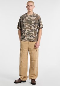 Camo t-shirt met korte mouwen in tinten van groen en bruin met een borstzak, gecombineerd met kaki cargo broeken met meerdere zakken. Bruine laarzen.