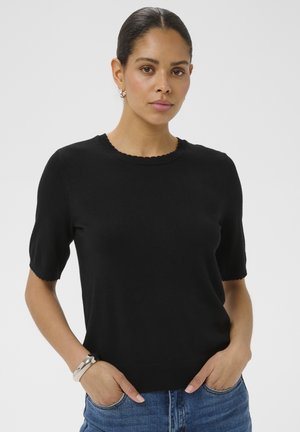 Femme aux cheveux foncés portant un pull noir à manches courtes et un jean bleu, debout les mains dans les poches devant un fond uni.