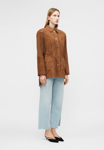 Γυναίκα που φοράει καφέ σακάκι από σουέντ με λεπτομέρειες με φουντίτσες, ανοιχτό μπλε τζιν παντελόνι με φαρδιά πόδια και μαύρες μυτερές γόβες με λουράκι, στέκεται πλαγίως.