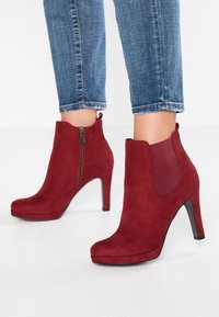 Red suede ankle boots with a high heel, featuring a side zipper and elastic panel, paired with blue denim jeans. Smooth texture.