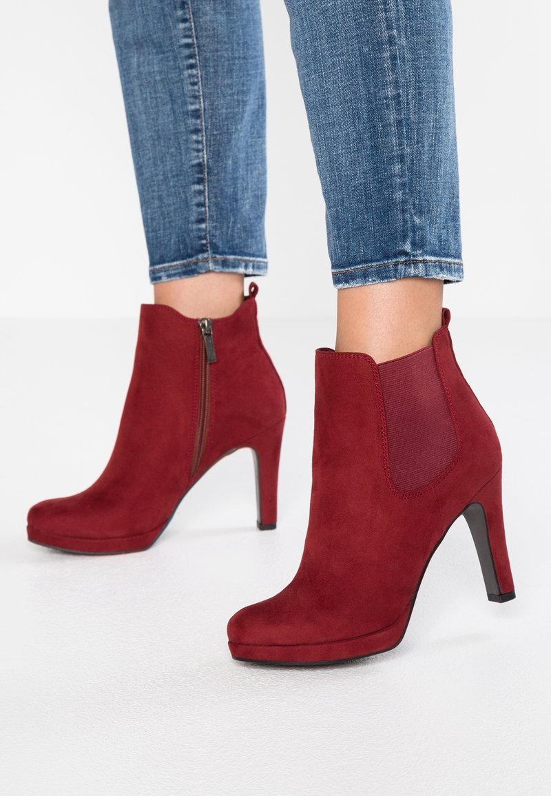 Red suede ankle boots with a high heel, featuring a side zipper and elastic panel, paired with blue denim jeans. Smooth texture.