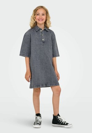 Enfant souriant avec des cheveux blonds bouclés portant une robe en denim grise ample et des baskets noires montantes, se tenant devant un fond uni.