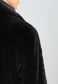 Svart fuskpälsjacka med en mjuk, texturerad finish och bred krage; bilden visar bakvyn med fokus på tyget och siluetten.