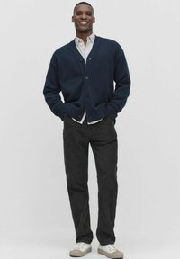 Mörkblå stickad cardigan med v-ringning och knappstängning, i kombination med en vit skjorta, svarta byxor och beige sneakers.