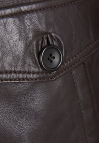 Brun læder med en struktureret overflade, der har en sort knap og kontrasterende syning langs sømmen. Nærbillede af knapdetaljen.