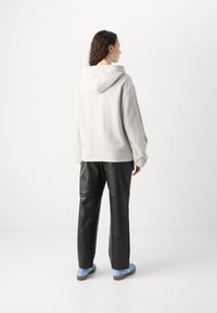 Felpa grigia chiara con tasca posteriore e cappuccio, abbinata a pantaloni di pelle nera e scarpe slip-on blu. Texture liscia, design casual.
