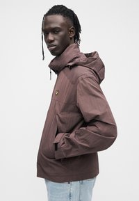 Giacca marrone leggera con cappuccio, dotata di una tasca con cerniera e un piccolo logo sul petto. Tessuto testurizzato con vestibilità rilassata.