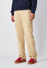 Pantaloni cargo beige con tasche laterali, taglio dritto, realizzati in morbido cotone. Abbinati a scarpe in suede rosse con accenti bianchi.