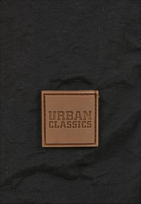 Brūns taisnstūrveida pleķis ar iegravētu tekstu "URBAN CLASSICS", novietots uz melna teksturēta auduma fona.