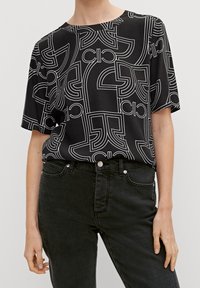 Sort kortærmet skjorte med et hvidt geometrisk mønster, løs pasform og rund halsudskæring, parret med sorte jeans.