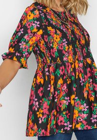 Top nero arricciato con stampa floreale brillante arancione, rosa e verde, maniche corte a volant, indossato con jeans blu e una collana d'oro.