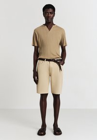Giovane uomo con pelle scura indossa una maglietta beige a righe con maniche corte, pantaloni corti beige con una cintura intrecciata marrone e sandali scuri intrecciati, in piedi.