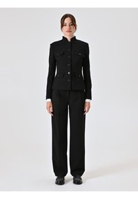 Zwarte getailleerde blazer met een hoge kraag, knoopsluiting en klepzakken, gecombineerd met loszittende zwarte broek en zwarte schoenen.