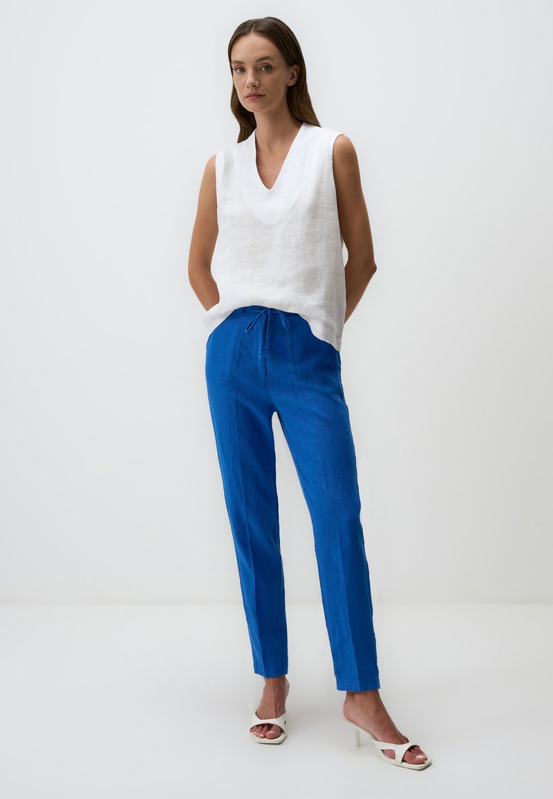 Witte linnen mouwloze top met een diepe V-hals, gecombineerd met blauwe linnen broek met een elastische tailleband en trekkoord. Gemaakt met witte hoge hakken sandaaltjes.