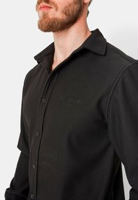 Zwarte geribbelde shirt met een kraag, voorzien van knoopsluiting en subtiele branding op de borst. Glad textuur van de stof zichtbaar.