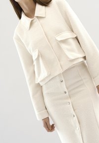 Valkoinen teksturoitu lyhyt takki kahdella etutaskulla, yhdistettynä yhteensopivaan polvenpituiseksi olevaan hameeseen, jossa on napit edessä.