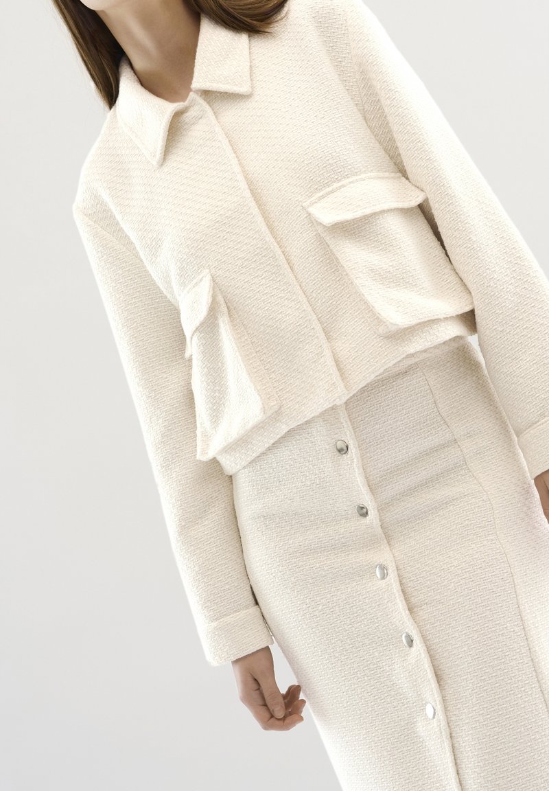 Valkoinen teksturoitu lyhyt takki kahdella etutaskulla, yhdistettynä yhteensopivaan polvenpituiseksi olevaan hameeseen, jossa on napit edessä.