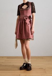 Femme portant une robe en satin rose poudré avec des manches en dentelle noire, des chaussettes beiges, des chaussures noires à boucle, et tenant une petite pochette dorée texturée.