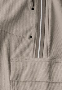 Beige rakenteellinen kangas, jossa on kaksinkertainen ommel, taskuläppä, metalliset nepparit ja ruskeavaihtoehtoisen ja valkoisen raidallisen vetoketjun yksityiskohta.