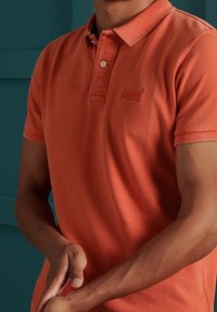 Polo orange avec col et patte de boutonnage à deux boutons ; matière en coton lisse ; petit logo sur la poitrine gauche ; manches courtes.