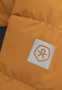 Orange vadderad tyg med synliga sömmar och en silverfärgad logotypatch med en cirkulär design och en abstrakt figur i mitten.