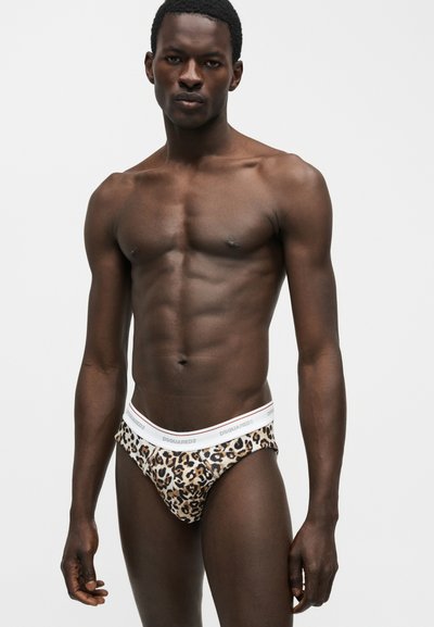 Λεπτός ανδρικός μοντέλο φοράει σλιπ με λεοπάρ μοτίβο και λευκή μέση με την επιγραφή "DSQUARED2" σε απλό ανοιχτό γκρι φόντο.