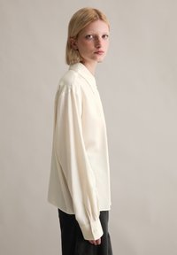 Μπλούζα σε ανοιχτό κρεμ χρώμα, από λεία ύφασμα, με κολάρο, μακριά μανίκια και κουμπωτές μανσέτες. Απλή, χαλαρή γραμμή.