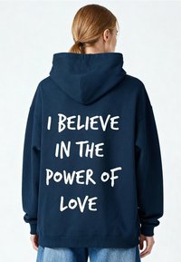 Sweat à capuche bleu marine à coupe ample, avec une poche kangourou et un texte blanc au dos indiquant "JE CROIS EN LA PUISSANCE DE L'AMOUR."
