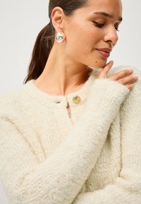 Cardigan en peluche couleur crème avec un col rond, fermeture à boutons et poignets côtelés. Le modèle porte des boucles d'oreilles métalliques et touche son épaule.