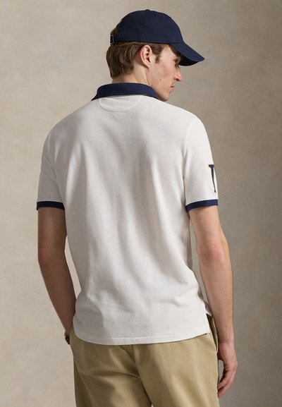 Λευκό πόλο πουκάμισο με ναυτικό γιακά και μανσέτες, με υφή υφάσματος. Φορεμένο με χακί παντελόνια και ναυτικό καπέλο.