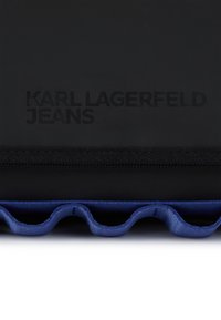 Μαύρη τσάντα με υφή, που διαθέτει κυματιστή μπλε λεπτομέρεια στη βάση. Το ανάγλυφο λογότυπο αναγράφει "KARL LAGERFELD JEANS" σε απαλό μαύρο.