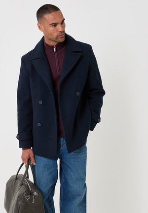 Mężczyzna w granatowym płaszczu pea coat i niebieskich dżinsach trzyma ciemnoszarą teksturowaną torebkę, patrząc w lewo na jednolitym tle.