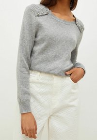 Pull en maille gris avec un col large, agrémenté de boutons argentés sur l'épaule, associé à un pantalon clair à pois.