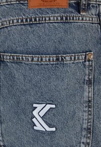 Denim džinsi gaiši zila krāsā, ar ādas uzšuvni un baltu izšūtu logotipu aizmugures kabatā.
