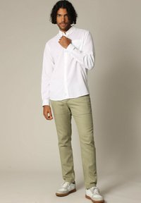 Chemise blanche à manches longues avec un poche poitrine, associée à un pantalon vert clair et des baskets blanches à semelles brunes. Texture de tissu lisse.
