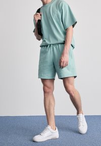 Vihreä minttukangas t-paita ja yhteensopivat lyhyet housut joustavalla vyötäröllä, valkoisten lenkkareiden kanssa, seisoo sinisellä matolla.