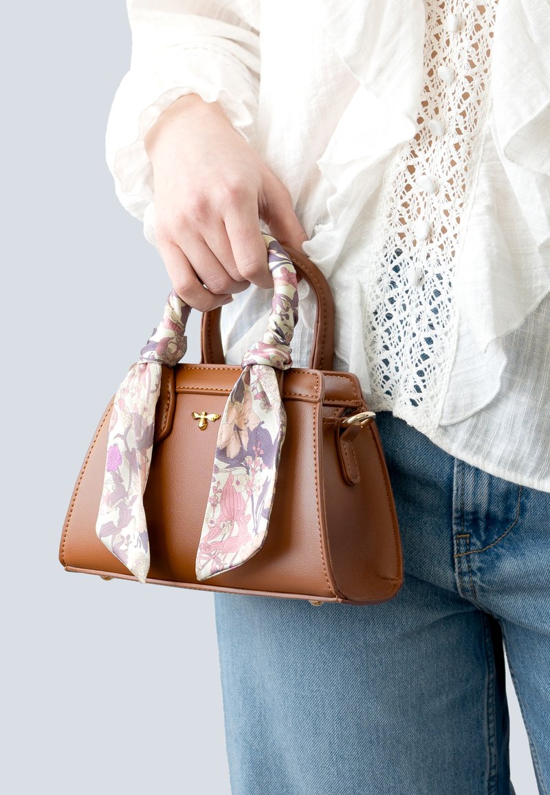 Brown leather handbag with a structured shape, featuring a decorative floral scarf tied around the handles. Smooth texture and gold hardware detail.