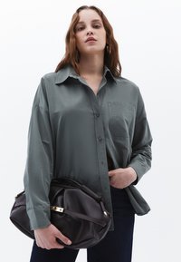 Camicia grigia a maniche lunghe con bottone e tasca sul petto, abbinata a una borsa in pelle marrone scuro con chiusura e zip dorate.