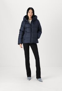 Mornarska puhasta jakna s kapuco, z vodoravno kviltanim dizajnom in sprednjim zadrgo. S kombinacijo črnih hlač in sivih čevljev z naostrenim prstom.