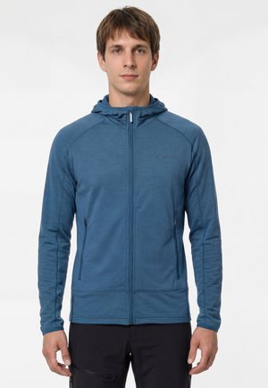 Jonge man draagt een blauw jack met rits en capuchon, met zakken aan de voorkant, staand tegen een effen lichte achtergrond.