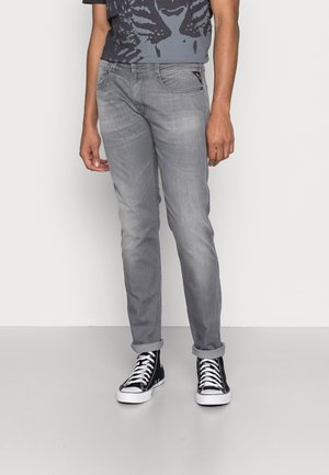 Mann trägt hochgekrempelte graue Jeans, schwarz-weiße High-Top-Sneaker und ein dunkles T-Shirt mit Grafik, steht vor einem schlichten weißen Hintergrund.