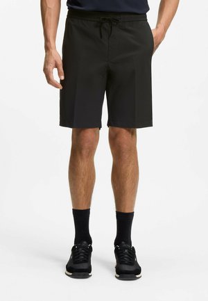 Mand iført sorte knælange shorts med snøre, sorte sokker og sorte sneakers, stående med hænderne nær lommerne på hvid baggrund.