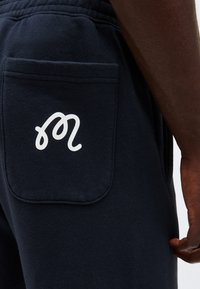 Close-up of a person wearing dark navy pants with a back pocket featuring a white stylized letter "m" and a hand resting nearby.